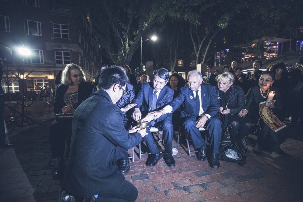 Guests seated at an evening outdoor gathering hold candles while a photographer kneels to take their picture